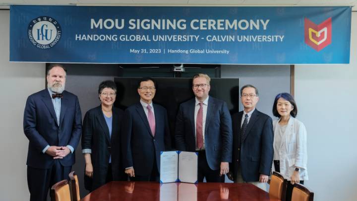 Four men and two women in suits standing and smiling with a signed agreement in the middle.