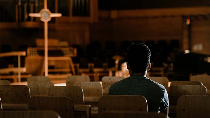 A person seated in a dark chapel with the cross illuminated in the front.