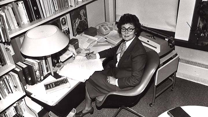 A black and white photo of a woman in a suit jacket and a skirt sitting at a desk near a bookshelf.