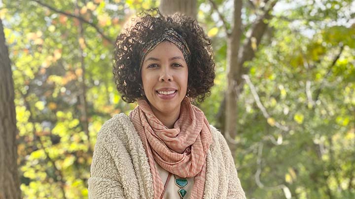 A woman smiling stands outside with trees in the background.