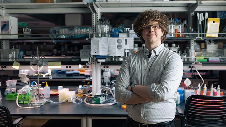 A college student with arms crossed stands in a science lab with equipment all around him.