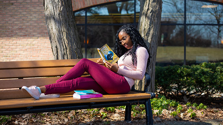 A student reads a book on a bench with a building in the background.