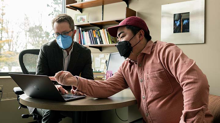 Two men wearing face coverings are seated in an office space reviewing data on a laptop computer.