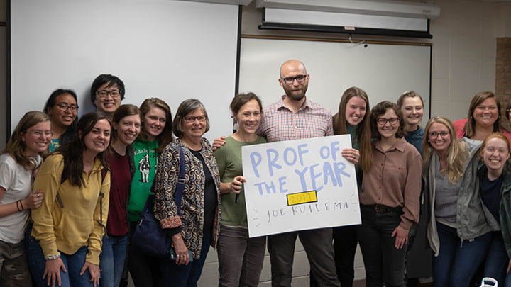 A professor holds a sign that says "Professor of the Year" surrounded by students.