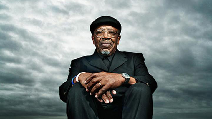 A man in a suit sits with his hands folded in front of a cloudy sky.