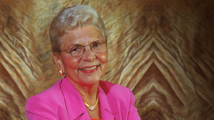 Headshot of a woman with gray hair in a pink blazer against a tan backdrop.