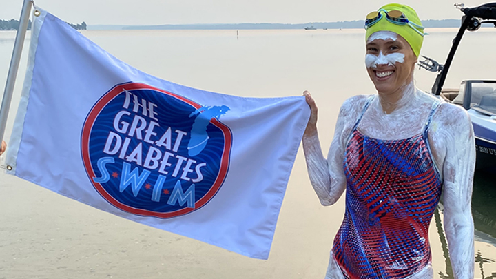 A woman in swimsuit, cap, and goggles holds "The Great Diabetes Swim" banner with a lake behind her.