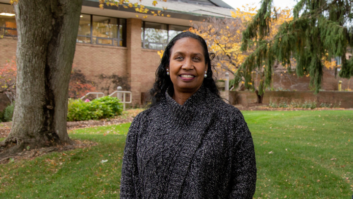 An African American woman smiling standing outside with a building and trees in the backdrop.