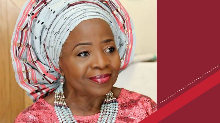 An African woman wearing a beautiful headdress, pink shirt, and pearl necklace.