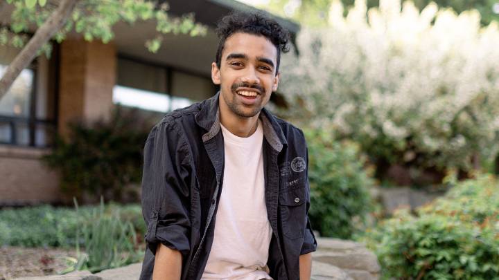 A student sits on a rock and smiles with a building and trees behind him.