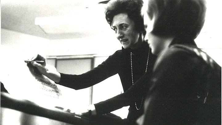 A female music professor standing next to and instructing a student who is seated at the piano.