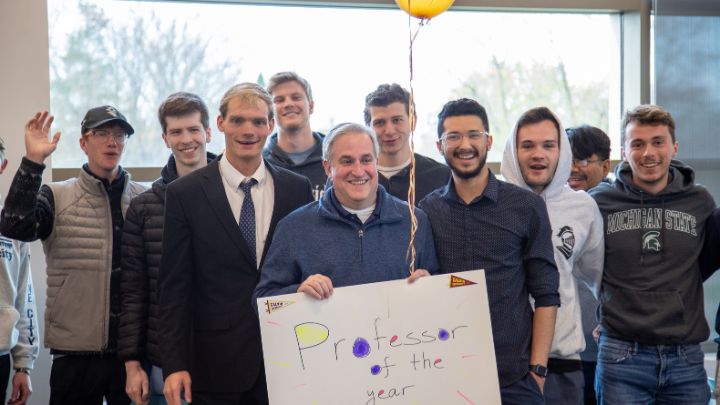 A man holding a sign saying "Professor of the Year" smiles with a group of students.