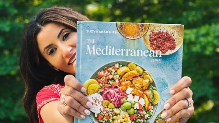 A woman smiling holds the cookbook she authored out in front of her toward the camera.