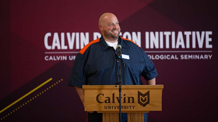 A man wearning prison blues smiles at a podium with a Calvin Prison Initiative banner behind him.