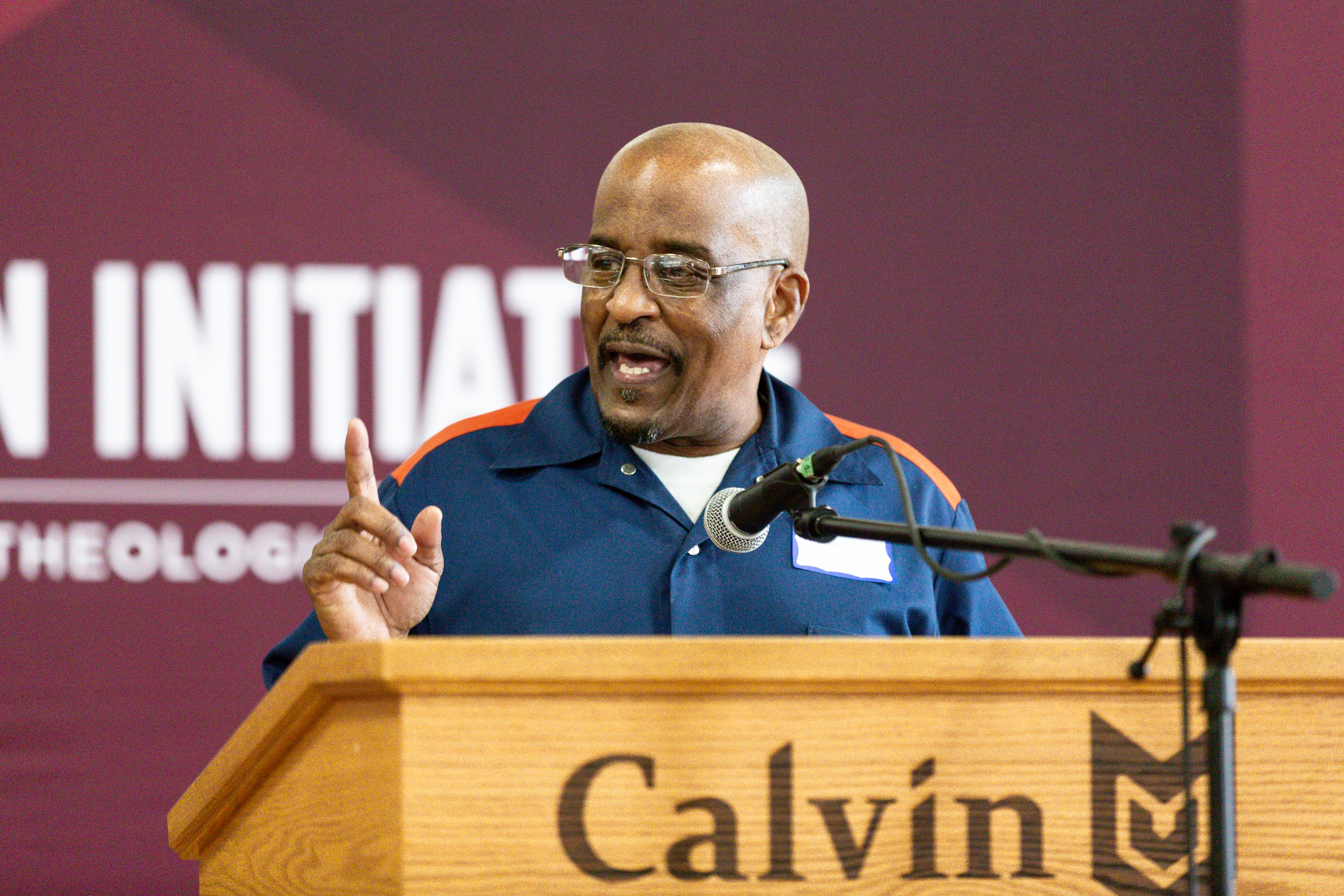 An African American man wearing glasses in prison blues speaks behind a podium.