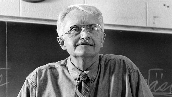 A black and white photo of an older professor with glasses in a classroom staring off to the right.