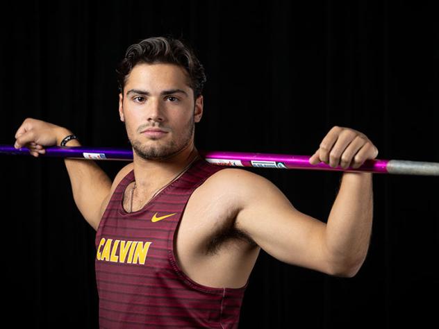 Colin Jambekar poses with a serious, competitive face, wearing a Calvin track and field jersey and holding a javelin.