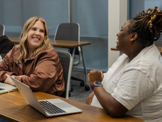 Two female students look at each other and smile. They are in a classroom with laptops open in front of each of them.