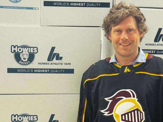 Max Sieplinga '04 standing with a Calvin hockey jersey on in front of Howie's HockeyTape boxes