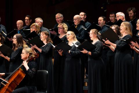 A choir performs with a cello on stage at Calvin University, dressed in black dresses and tuxedos, holding folders.