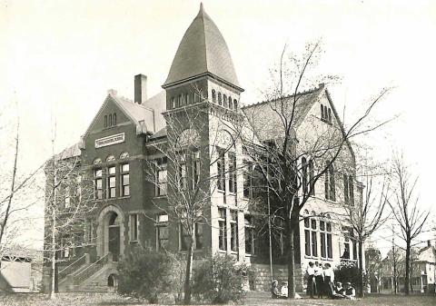 An old photo of a large schoolhouse, with students sitting and standing on the lawn in front.