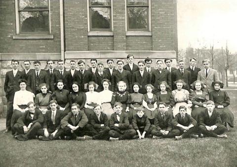 An old photo of a group of male and female students, wearing dresses, suits and ties, pose for a class photo.