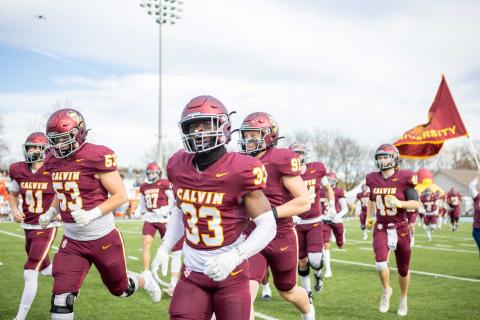 The Calvin University football team, wearing home maroon jerseys, run onto the field with the Calvin flag flying behind.
