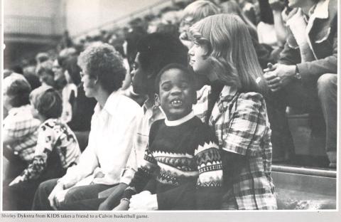 A historical photo of Calvin students with the children they mentor through the KIDS program, sitting in the stands at a basketball game.
