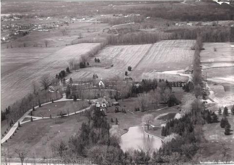 A historical photo of farmland from the air, with a couple homes, driveway, and a pond.