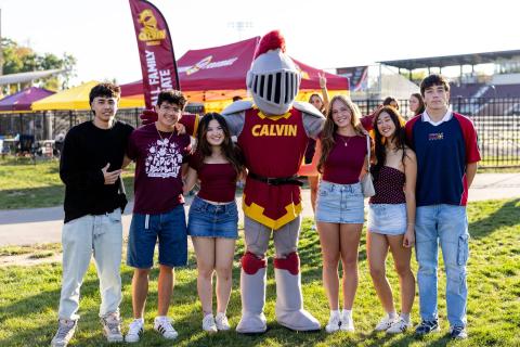 A group of students in maroon and gold stand with Joust the Calvin Knights mascot at a tailgate with tents in a field in the background.