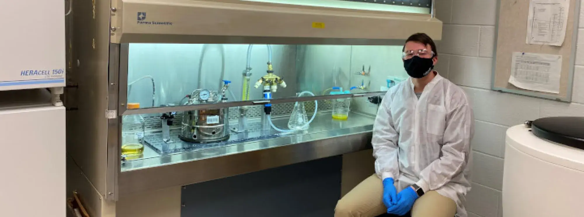 A research assistant sits next to his experiment that he's conducting within a laboratory.