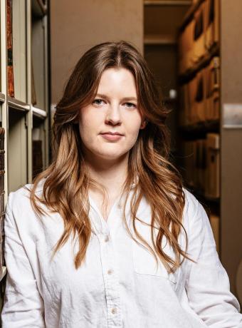 Jen Vos with long caramel brown hair in a white button down shirt leaning against a bookshelf in a library