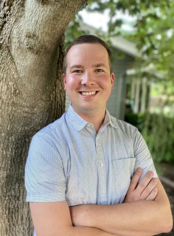 Caleb Lagerwey in a white short sleeved button down shirt leaning against a tree with his arms crossed