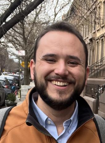 Close up photo of Isaac LaGrand smiling with leafless trees on a city street behind him