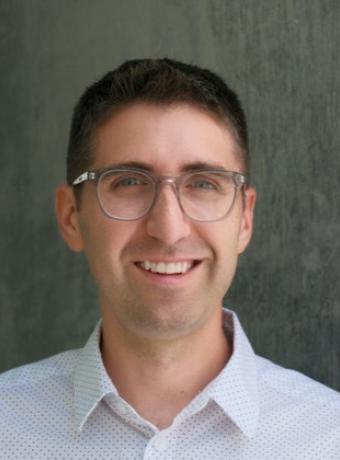 Dan Paulson in a white collared shirt with small black polka dots in front of a gray background