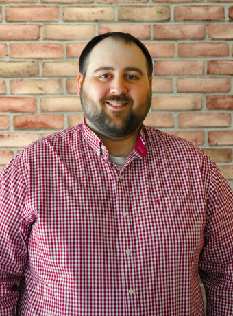 Man with dark hair and facial hair standing in front of a brick wall.
