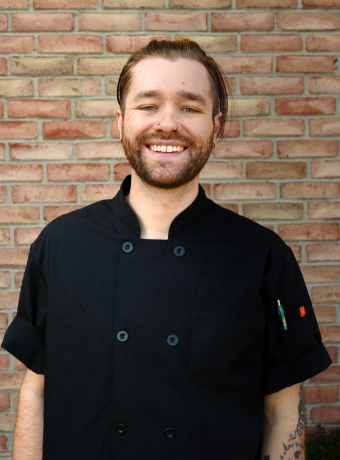 Man with facial hair wearing a black chef's coat, standing in front of a brick wall.