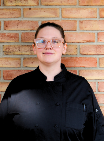 Woman with pink glasses wearing a chef's coat, standing in front of a brick wall.