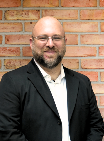 Man with glasses and facial hair wearing a black jacket, standing in front of a brick wall.