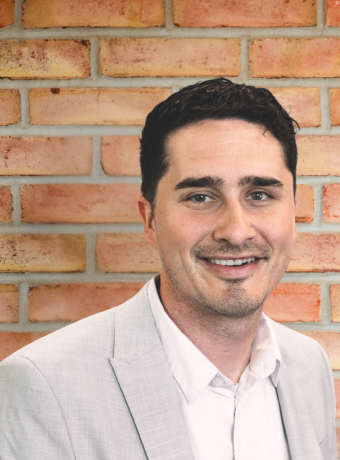 Man with black hair standing in front of a brick wall. 