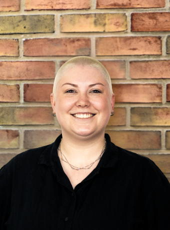 Woman in black shirt with blonde shaved head smiling in front of a brick wall.