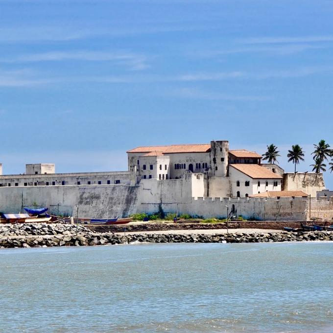 Elmina Castle under a blue sky, surrounded by palm trees.