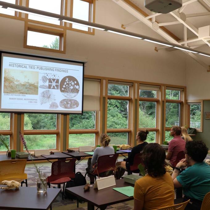 Someone leads a lecture for students in a room with high vaulted ceilings and a wall of windows looking out over green woods.