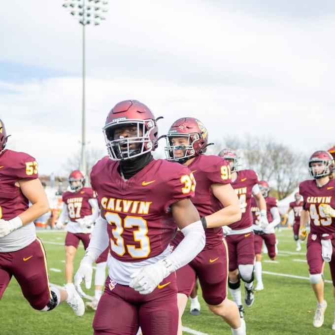 The Calvin University football team, wearing home maroon jerseys, run onto the field with the Calvin flag flying behind.