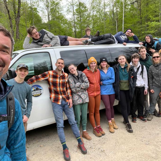 A group of staff and students in front of a 15 passenger van, wearing outdoor gear in the woods.