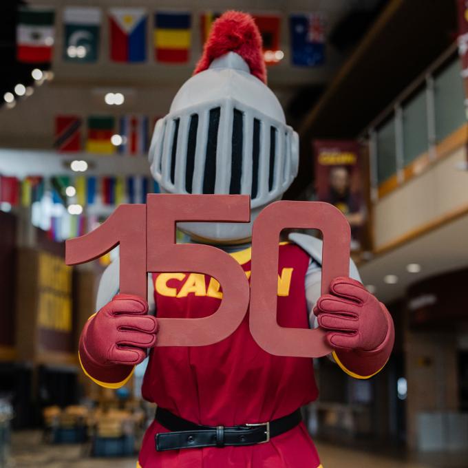 Joust the Calvin Knights mascot hold the number 150 in block letters in the Spoelhof Fieldhouse lobby.