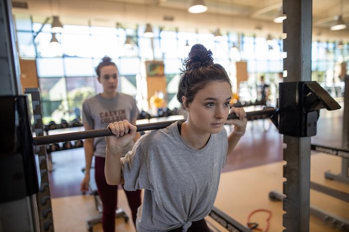 A female student in a gray t-shirt stands with the Smith machine barbell in the Calvin fitness center, with another student looking on.