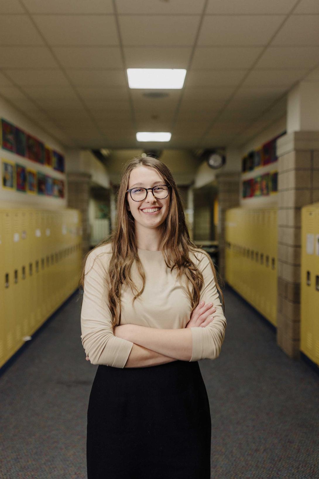 A student teacher folder her arms and smiles in a hallway of lockers.