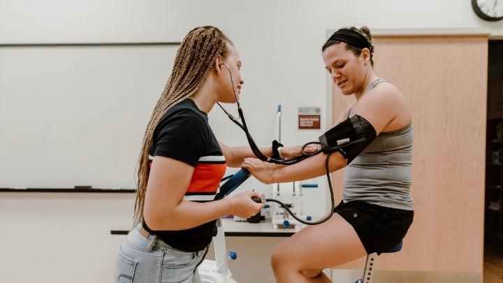 A women monitors the blood pressure of another student who's sitting on a stationary exercise bike.