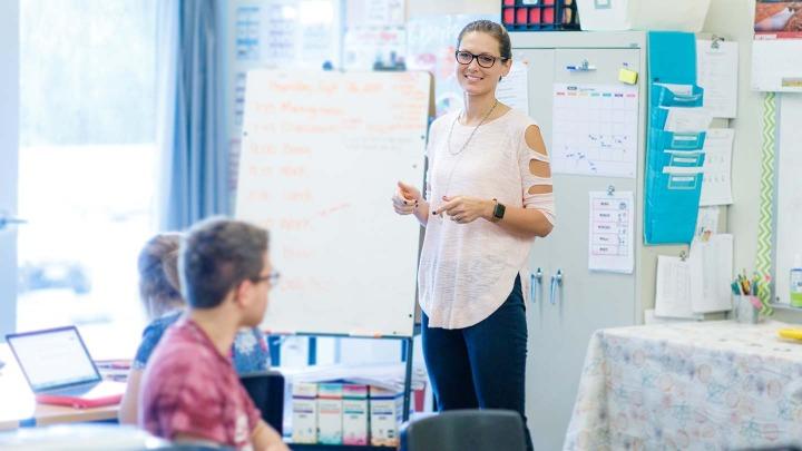 A teacher stands next to a whiteboard in front of a classroom of students.